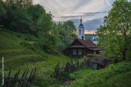 Beautiful spring landscape overlooking an old wooden house. In the background is a view of the bell tower and domes of the Varvarinskaya Church, amazing sunset in the city of Ples.