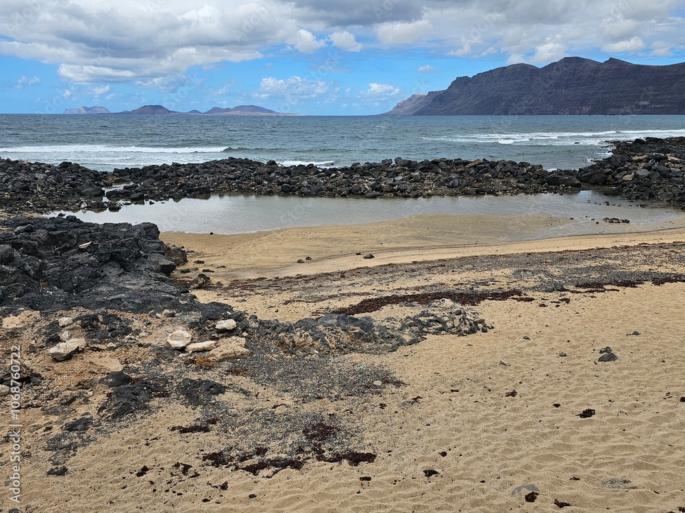 Une plage de sable avec des formations rocheuses volcaniques et des flaques d'eau, face à l'océan et encadrée par des montagnes, avec des îles visibles au loin sous un ciel nuageux. Une plage de sable avec des formations rocheuses volcaniques et des flaques d'eau, face à l'océan et encadrée par des montagnes, avec des îles visibles au loin sous un ciel nuageux.