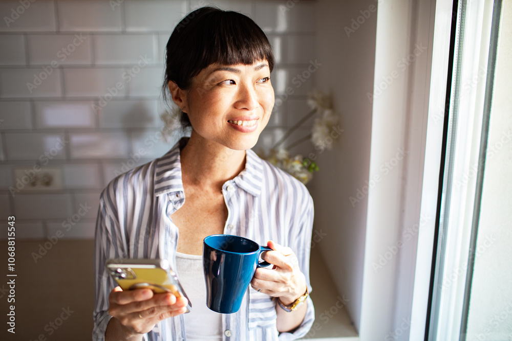 Woman smiling while checking her phone and holding a coffee mug in a bright kitchen Woman smiling while checking her phone and holding a coffee mug in a bright kitchen