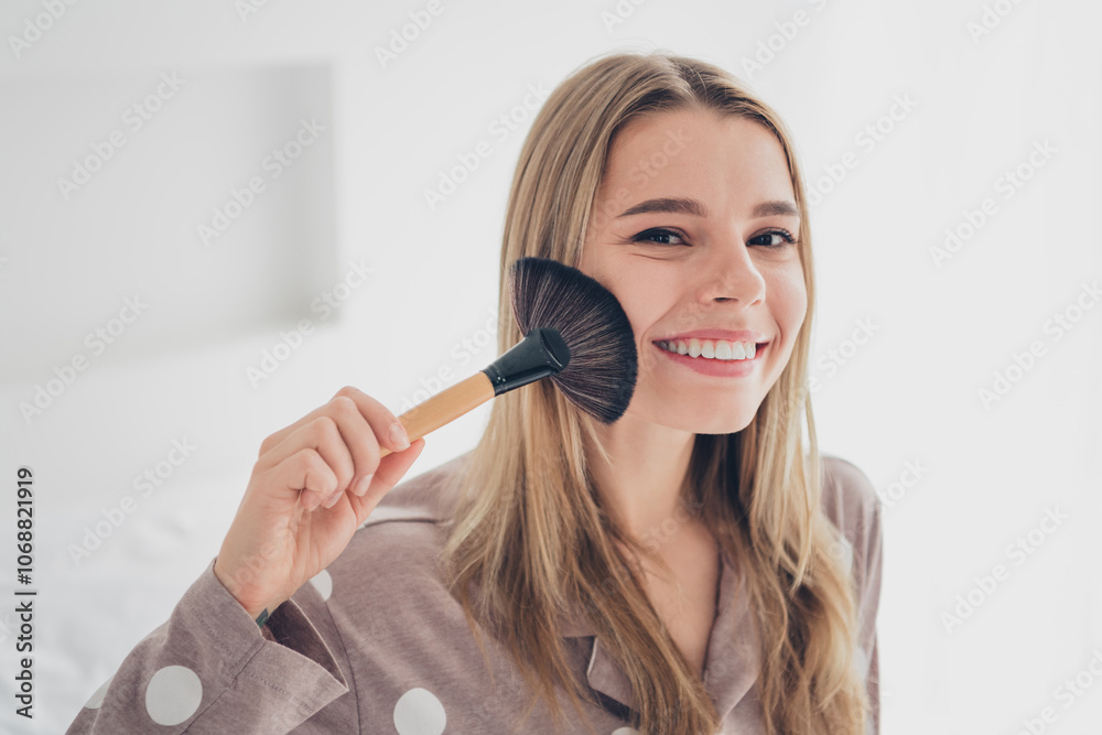 Young woman smiling in bedroom wearing pajamas and applying makeup with brush in calming natural light Young woman smiling in bedroom wearing pajamas and applying makeup with brush in calming natural light