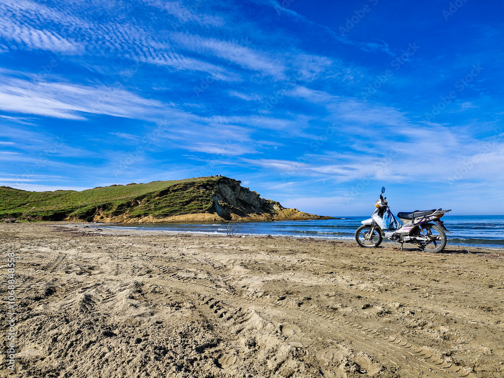 Fototapeta premium A motorcycle standing alone on the shore of the sea on the beach of Zvernec in Albania, in the foreground are tire tracks in the sand, in the background is the sea surface and rocks with trees and bus