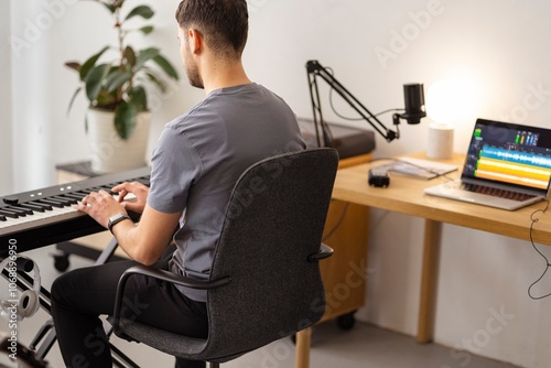 A man sits focused at a keyboard in a minimalist home studio setup, with a laptop showing audio tracks on the desk nearby, creating a dedicated space for sound editing and music production photo