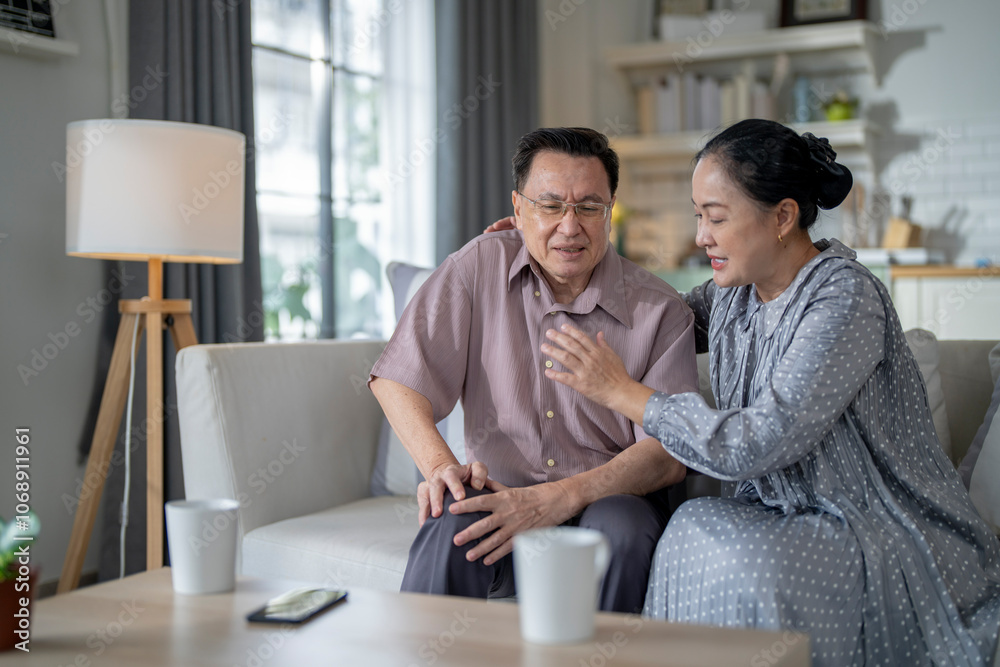 An elderly couple sits in their living room. The man holds his knee, appearing to be in discomfort, while his partner offers support. The image conveys care, concern, and the bond between the couple. An elderly couple sits in their living room. The man holds his knee, appearing to be in discomfort, while his partner offers support. The image conveys care, concern, and the bond between the couple.