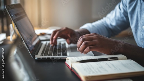A man is typing on a laptop in front of a notebook and a cell phone. The laptop is open and the man's hand is on the keyboard. The scene suggests that the man is working or studying