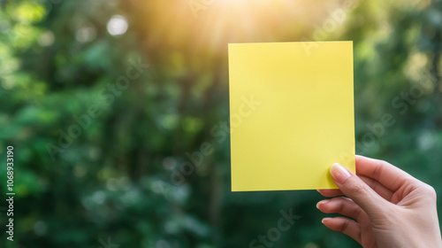 hand holding yellow sticky note against blurred green background, symbolizing creativity and ideas. warm sunlight adds positive vibe photo