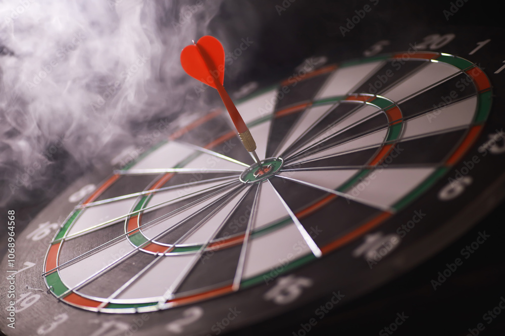 Fototapeta premium Red Dart Hitting Bullseye on Traditional Dartboard with Smoky Background