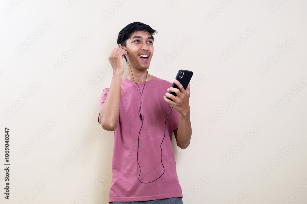 Asian man in casual pink t-shirt wearing earphones listening to music from smartphone isolated on cream background