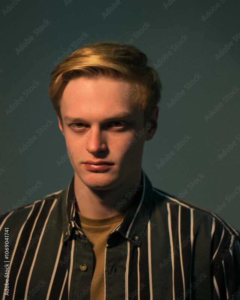 Portrait of young man with blond hair and blue eyes, wearing striped shirt, isolated on dark background, looking straight at camera Portrait of young man with blond hair and blue eyes, wearing striped shirt, isolated on dark background, looking straight at camera