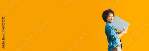 A young curious man with curly hair, wearing a blue denim shirt, stands against a vibrant orange background while holding a laptop close to his chest. Ample copy space on the left side