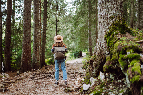 Woman in hat with backpack walks along ground road in dense forest. Fresh air and wild nature. Hiking.