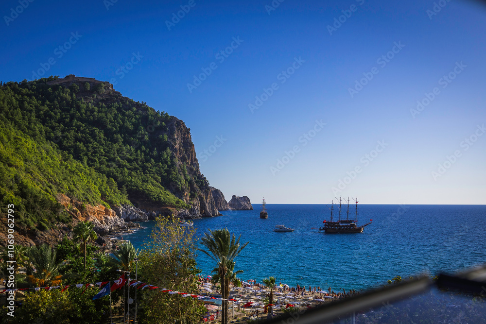 Fototapeta premium Alanya castle top view on the mountain with coast ferry boat on blue sea . Beautiful cleopatra beach Alanya Turkey landscape travel landmark.