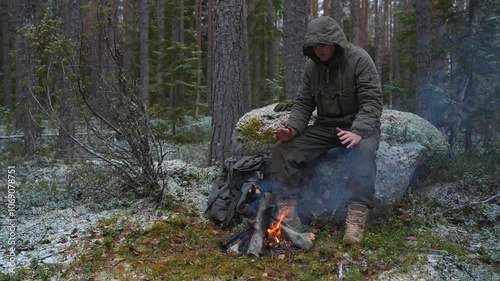 Hooded Camper by the Fire in a Snowy Landscape. Forest man warming by the fire in the frosty forest. The man in the hood sits by the fire. Forester man warms his hands near the campfire