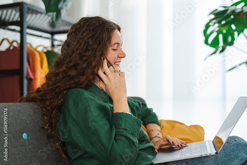 Young woman multitasking by talking on her phone and using a laptop while relaxing on a couch in a comfortable home environment photo