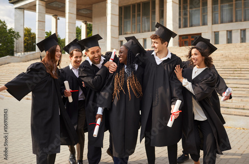 Group of a smiling happy multiracial excited graduates students hugging and having fun in a university graduate gown holding diploma in hands outdoor. Education and graduation concept.