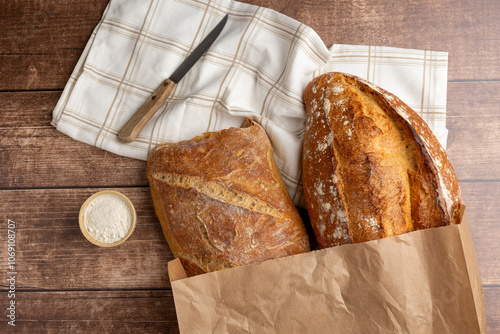 Freshly baked artisanal bread in a paper bag on a wooden table, accompanied by a rolling pin and a small bowl of flour, ready for a perfect breakfast.