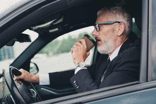 Mature businessman in formal suit enjoying coffee while driving car in city street