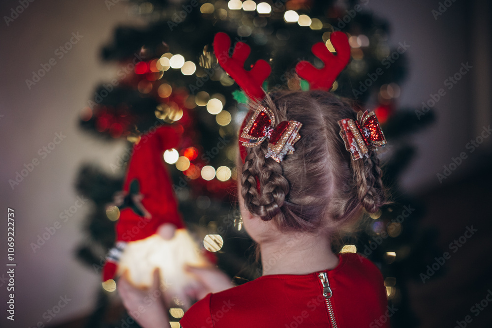 Naklejka premium Blonde girl in a beautiful red dress with a Christmas hairstyle and beautiful hairpins on her head against the background of a Christmas tree