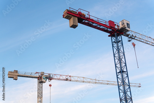 A pair of tower cranes rises into the sky, adding an urban accent against the clear blue background. The simplicity of the forms and the minimalist composition emphasize their structure. photo