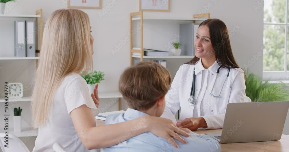 Satisfied mother and her little son have fun talking to pediatrician during visit to hospital. Woman puts reassuring hand on boys back, showing positive and supportive atmosphere during meeting. Satisfied mother and her little son have fun talking to pediatrician during visit to hospital. Woman puts reassuring hand on boys back, showing positive and supportive atmosphere during meeting.
