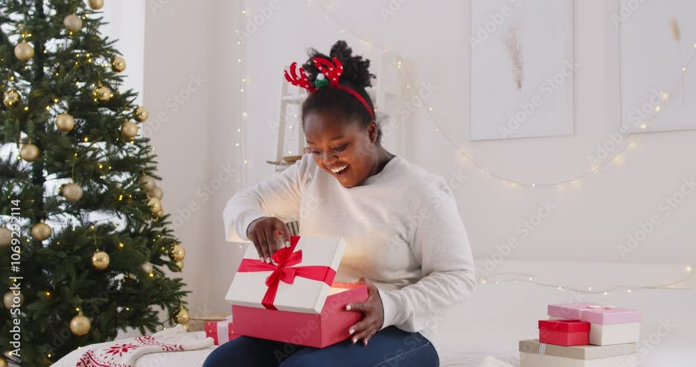 African American woman celebrates Christmas or New Year by opening a gift box at home. The holiday highlights excitement and surprise as she unwraps the present in a joyful atmosphere.