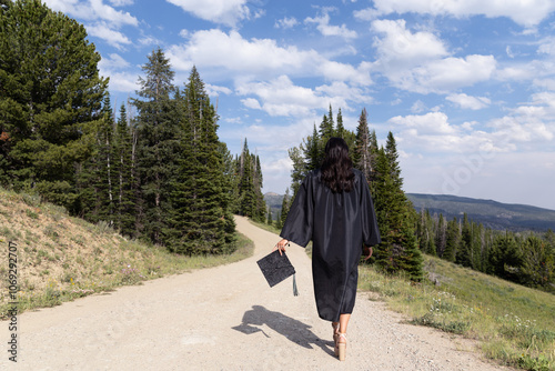 Teenage girl in high school graduation cap and gown walking away on a dirt road in the mountains with her back to the camera to illustrate walking into future to start new chapter in life graduating