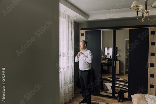 A man is getting dressed in a room with a window and a mirror. He is wearing a suit and tie