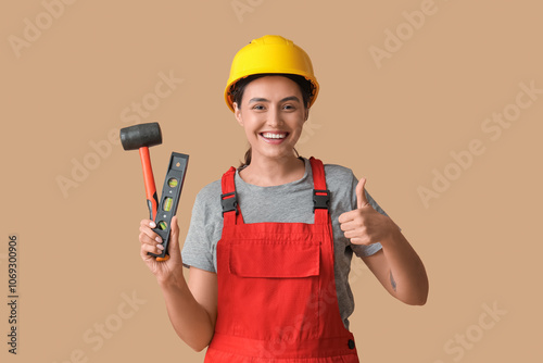 Young female construction worker with rubber mallet and bubble level showing thumb-up on beige background. Woman in male profession concept photo