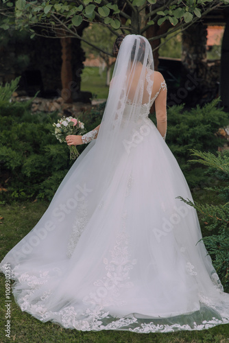 A woman in a white wedding dress is holding a bouquet of flowers. She is standing in a grassy area with trees in the background