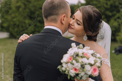 A man and woman are kissing in a park. The man is wearing a suit and the woman is wearing a wedding dress. They are holding a bouquet of flowers