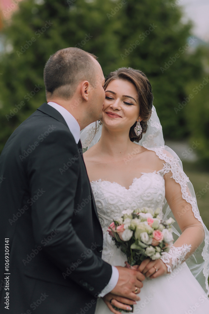 A man kisses a woman's cheek. The woman is wearing a wedding dress and holding a bouquet of flowers A man kisses a woman's cheek. The woman is wearing a wedding dress and holding a bouquet of flowers