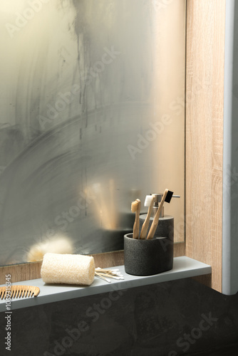 Neat bathroom shelf with wooden toothbrushes in a black container, a white towel, wooden comb, and cotton swabs, set against a foggy mirror. Emphasizes a well-organized and eco-friendly bathroom