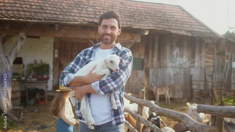 Handsome Caucasian male holding small white goat with both hands. Bugging beloved animal. Looking directly at camera while smiling. Other animals pulling their heads through wooden fence. Handsome Caucasian male holding small white goat with both hands. Bugging beloved animal. Looking directly at camera while smiling. Other animals pulling their heads through wooden fence.