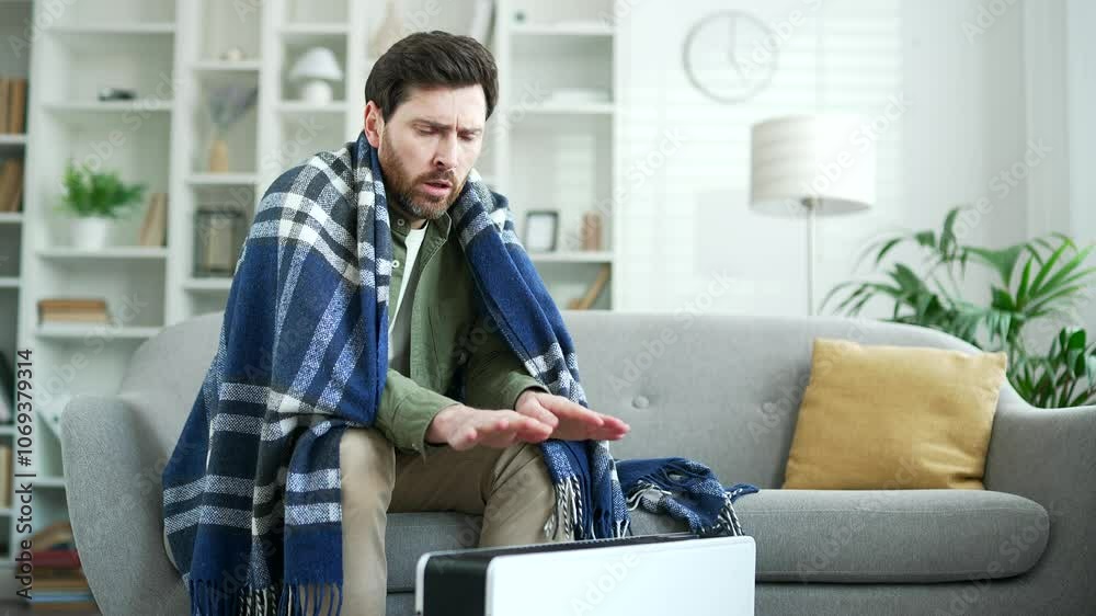 Upset man covered with a blanket frozen sitting on sofa in living room at home. Disappointed bearded mature male cold indoors and warms his hands to the heater, feels discomfort due to low temperature