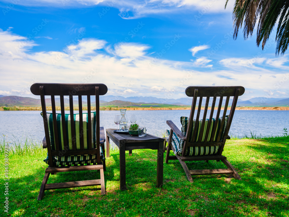 Lakeside scene with two wooden chairs under a palm tree, clear blue sky and white clouds, Robertson, Western Cape, South Africa
