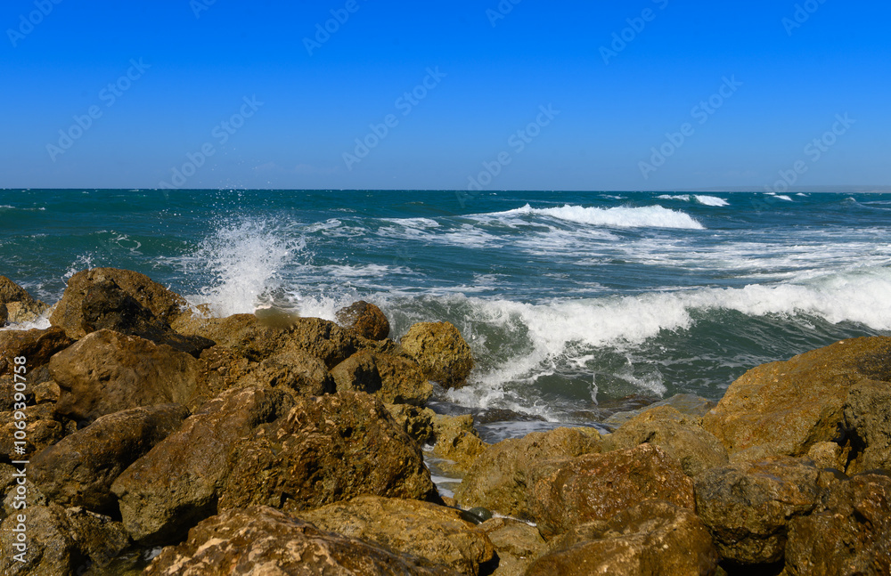 Waves crashing against rugged rocks under a clear blue sky by the seaside at midday in a coastal paradise Waves crashing against rugged rocks under a clear blue sky by the seaside at midday in a coastal paradise