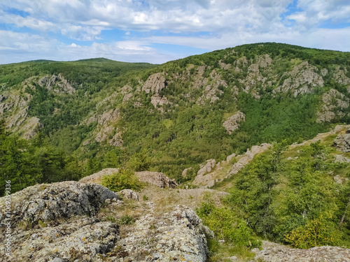 A majestic mountain, with vibrant trees and rugged surfaces, contrasts beautifully against a clear blue sky with fluffy clouds, creating a serene and inspiring natural scene