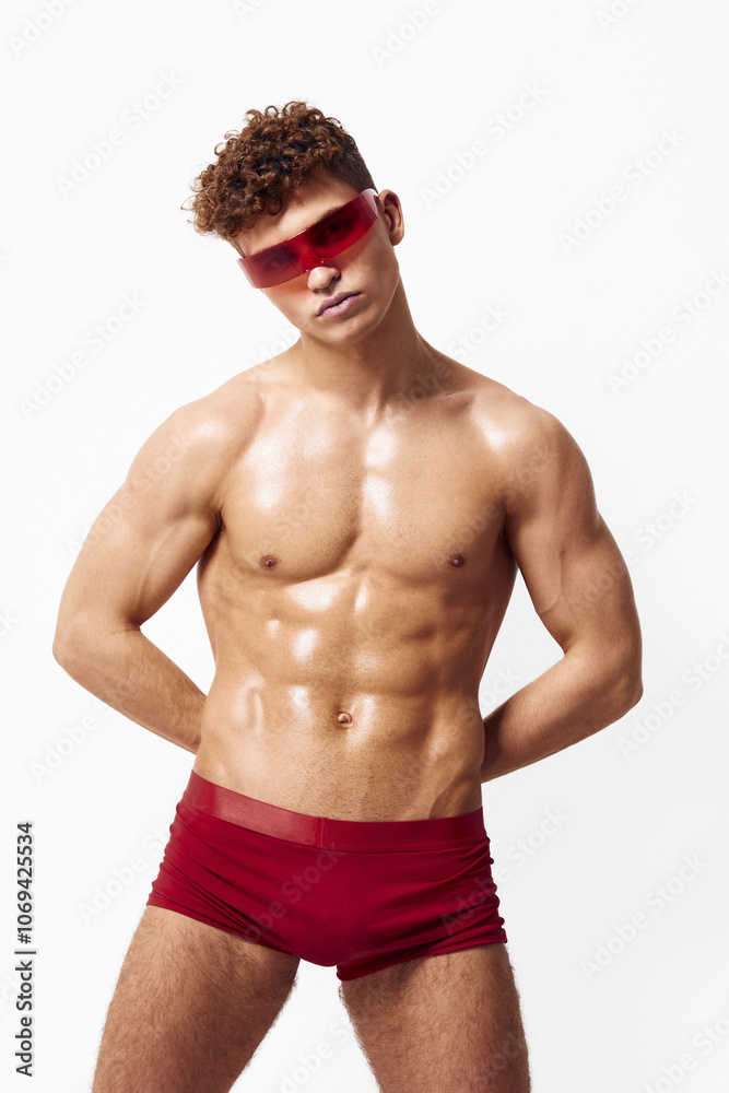 Fototapeta premium Confident young man posing in stylish red sunglasses and fitted swimwear, showcasing a toned physique against a clean white background. Fashion and summer lifestyle concept.