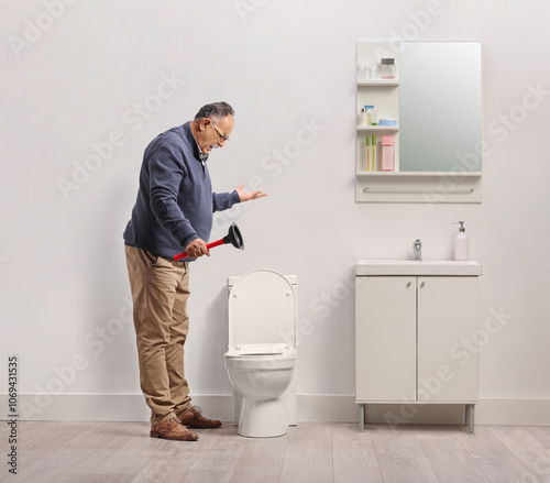 Fotografie Angry man holding a vacuum plunger and looking at a clogged toilet