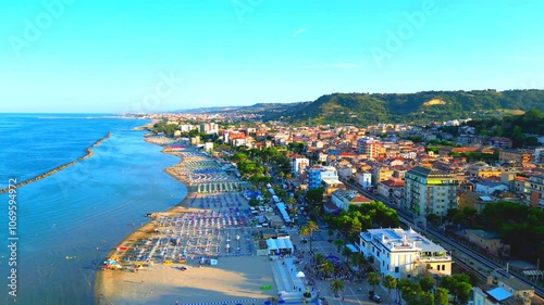 Stunning aerial truck left and pan right shot from Grottammare, with Adriatic Sea waters, sandy beach, a lot of parasols, buildings, humans, palm trees, railway, extending to far-away green hillscapes photo