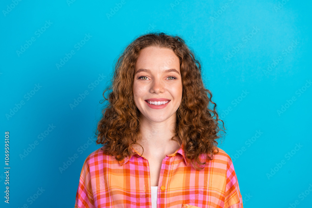 Fototapeta premium Cheerful young woman with curly hair in a plaid shirt, standing against a blue background, smiling warmly at the camera.