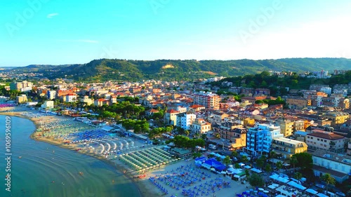 Luminous aerial push-in shot from Grottammare with golden hour sunlight permeating the manifold buildings, parasols, bathers, trees, the balmy waves of the Adriatic Sea, the distant green hillscapes photo