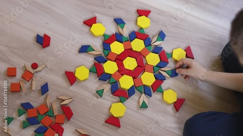 A European boy lays out a circular mandala pattern from wooden geometric figures. A child is concentrating on assembling a flower