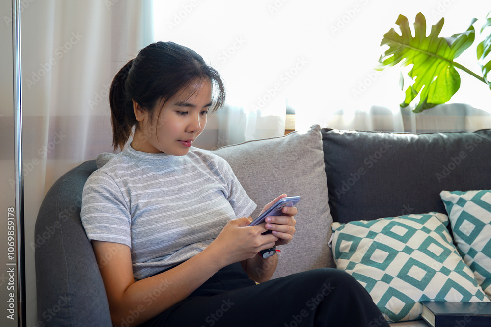 Asian woman sitting on sofa in living room chatting with friends with application in mobile phone in concept of leisure activity, relax, chilling, vacation. Soft and selective focus. Asian woman sitting on sofa in living room chatting with friends with application in mobile phone in concept of leisure activity, relax, chilling, vacation. Soft and selective focus.