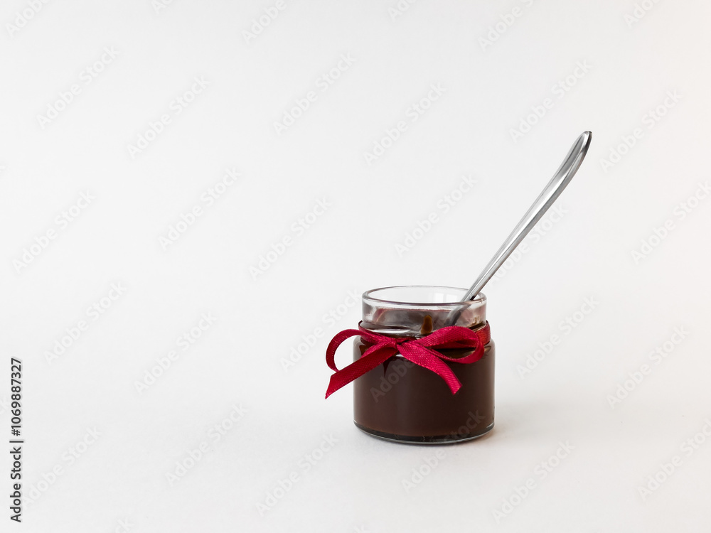 Small glass jar of chocolate spread with red ribbon and spoon on white background Small glass jar of chocolate spread with red ribbon and spoon on white background