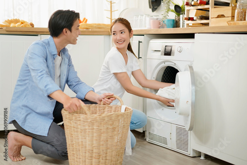 Happy young asian couple doing laundry fold clothes together using a front loading washing machine to wash laundry. Husband and wife during household routine in morning at home.