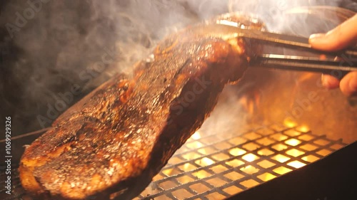 Close-up of meat grilled on barbecue over an open fire with herbs and rosemary. cook turns over toasted piece of meat with tongs. picnic in summer. cooking meat barbecue steaks in flames and smoke