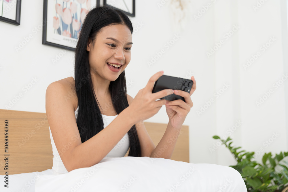Cheerful young Asian woman playing games on mobile phone on bed in the bedroom Cheerful young Asian woman playing games on mobile phone on bed in the bedroom