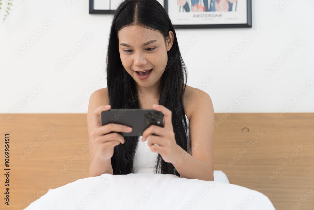 Cheerful young Asian woman playing games on mobile phone on bed in the bedroom Cheerful young Asian woman playing games on mobile phone on bed in the bedroom