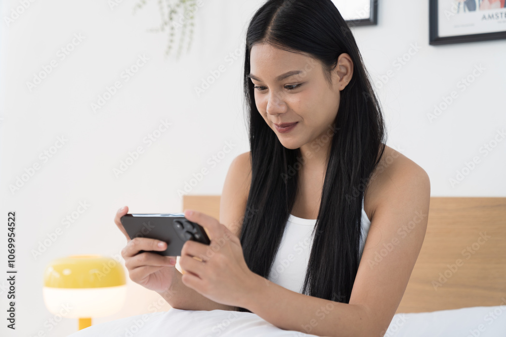 Cheerful young Asian woman playing games on mobile phone on bed in the bedroom Cheerful young Asian woman playing games on mobile phone on bed in the bedroom