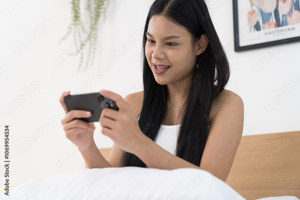 Cheerful young Asian woman playing games on mobile phone on bed in the bedroom